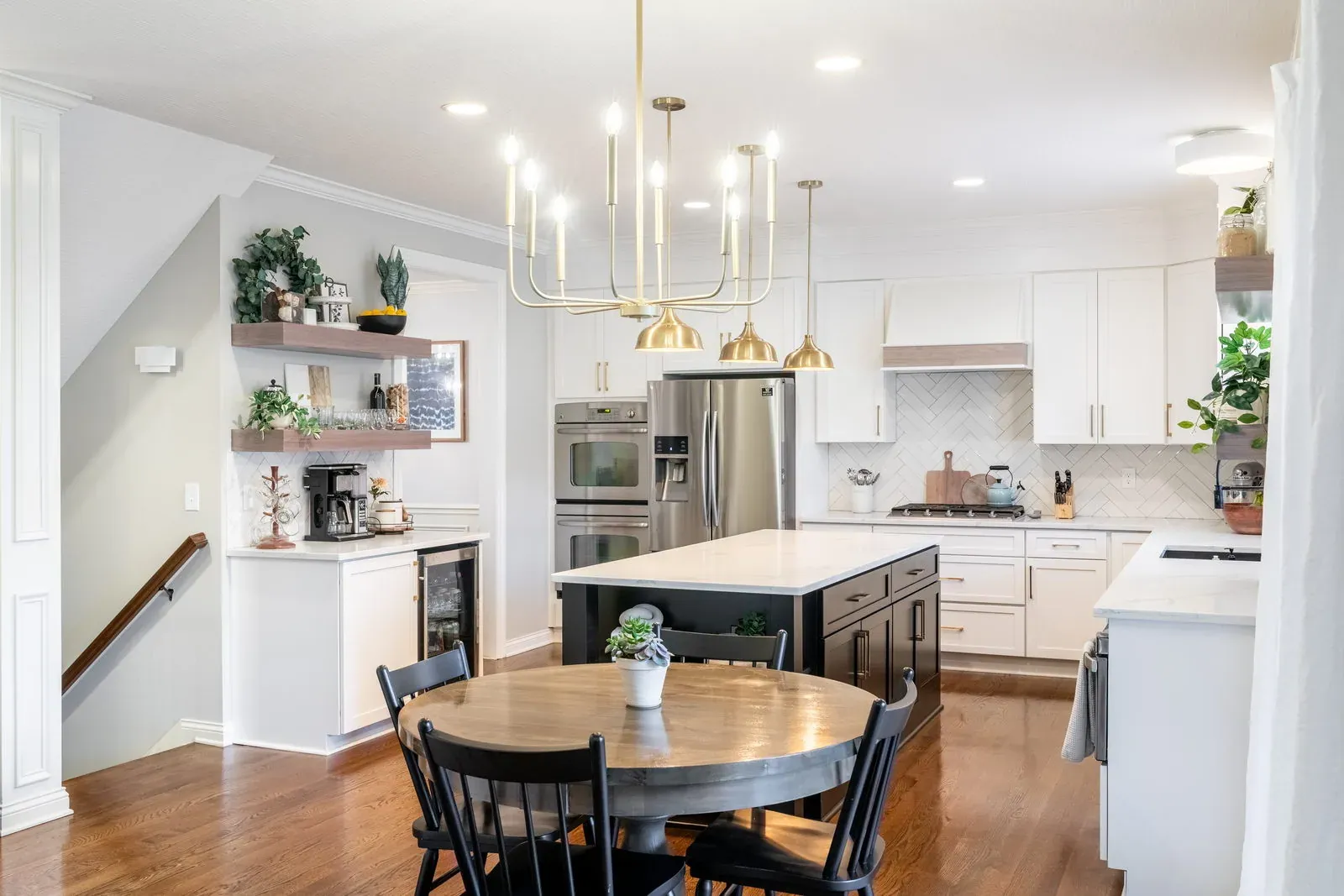 The same kitchen after cabinet refacing, with white shaker doors, a black island, quartz countertops and brass pendants over a round dining table