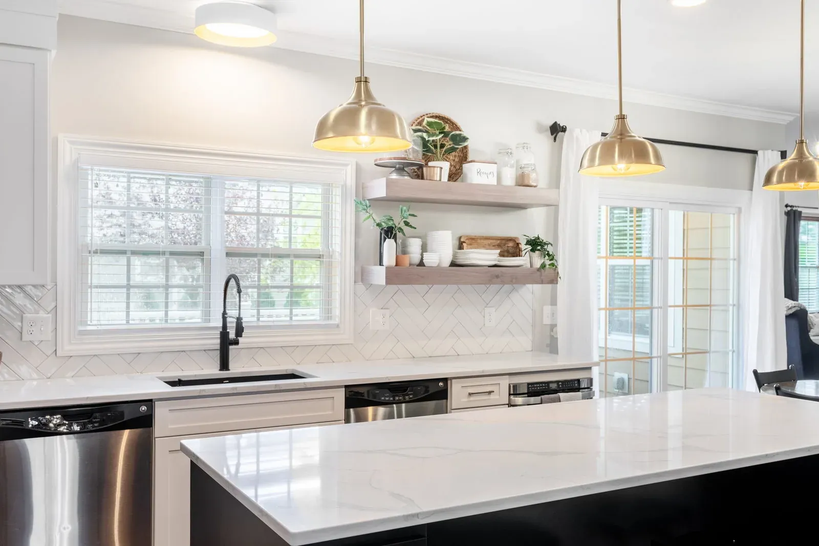 The same sink wall after refacing, with white shaker fronts, quartz countertops, herringbone tile, a matte black faucet and stained floating shelves