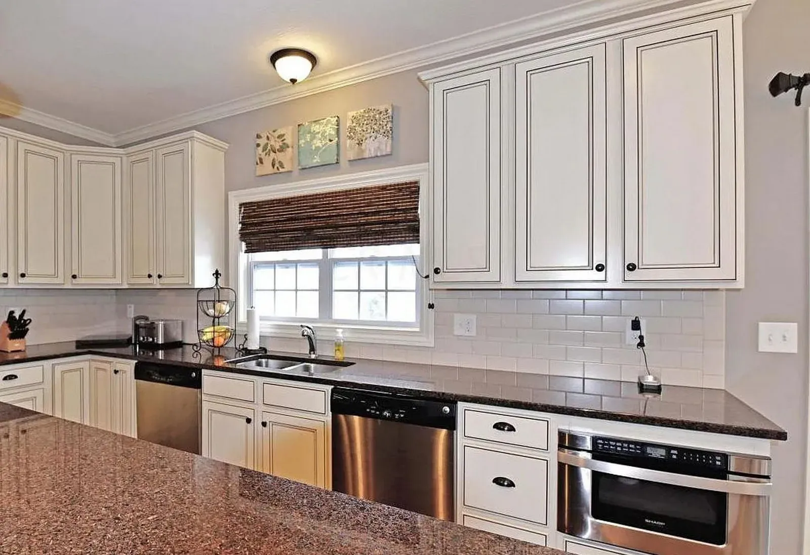 Kitchen sink wall before refacing, with glazed cream cabinets, speckled granite countertops and a white subway tile backsplash under a bamboo shade