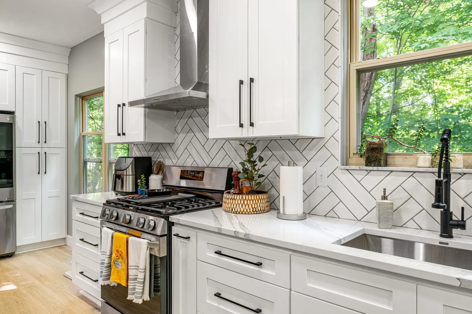 Stainless gas range set into white shaker cabinets with black bar pulls, quartz countertops and a herringbone tile backsplash