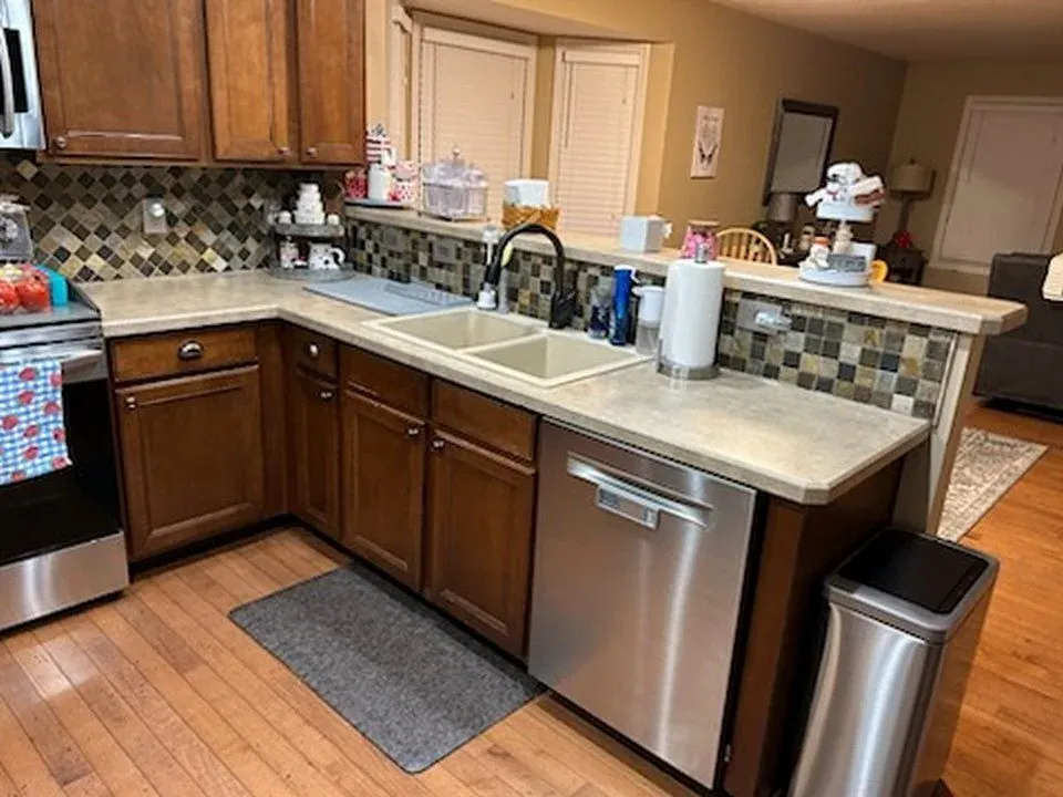 L-shaped kitchen before refacing, with dark cherry cabinets, a double-bowl sink, laminate countertops and a mosaic tile backsplash