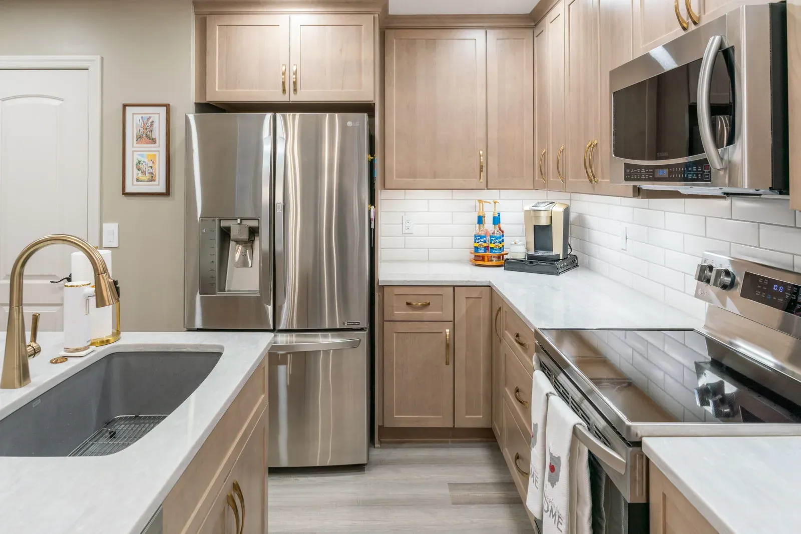 Refaced natural maple cabinets with a quartz counter, undermount sink, brass faucet and white subway tile beside a stainless refrigerator