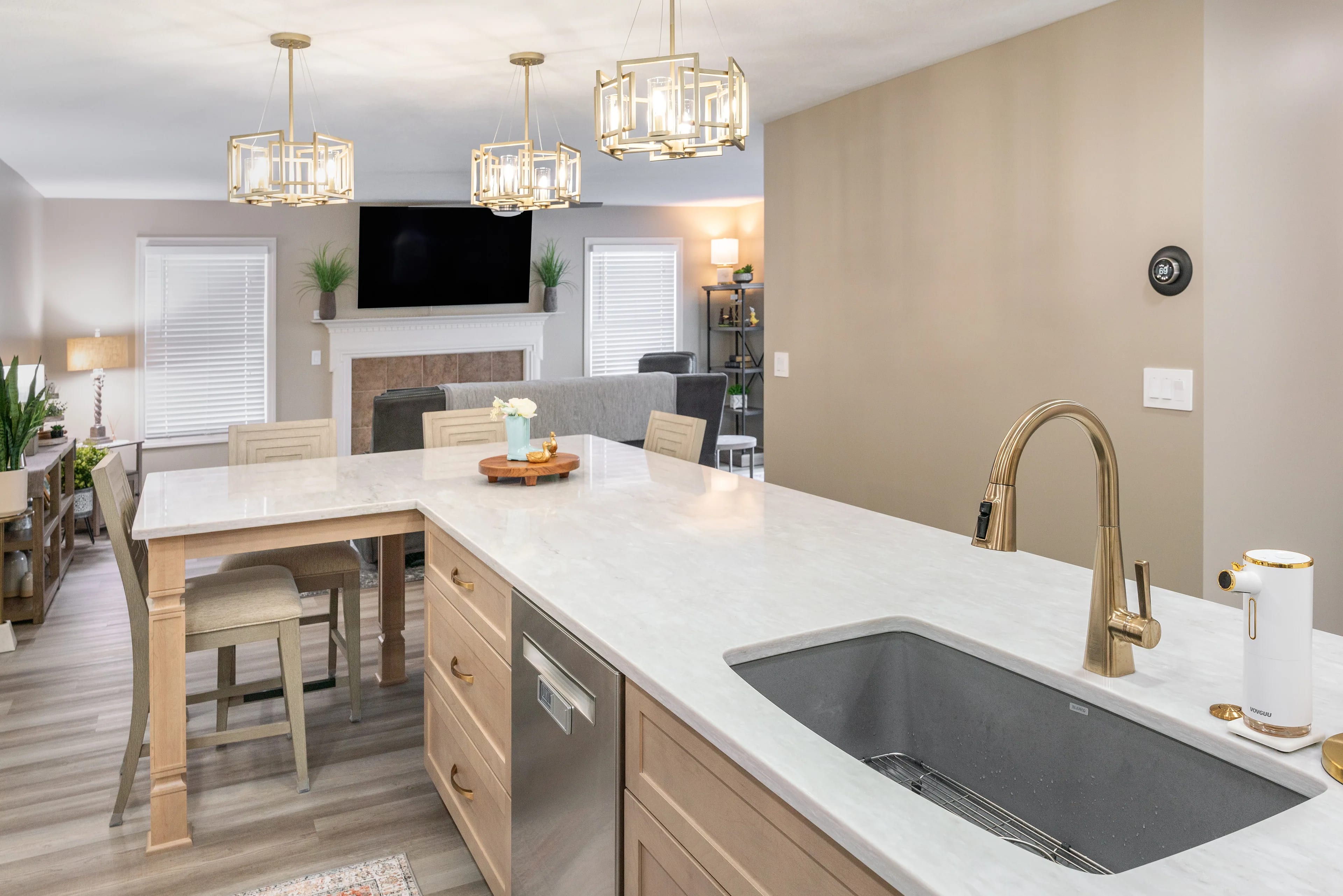 Refaced natural maple kitchen cabinets with quartz counters and a built-in dining table, looking through to a lamp-lit family room with a fireplace