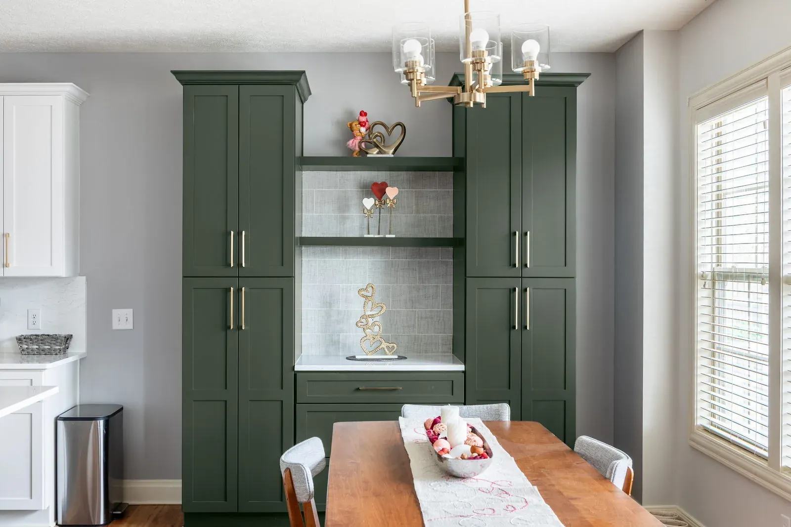 Custom hunter green dining hutch with glass-tile back panel and open shelving beside a walnut dining table under a brass chandelier
