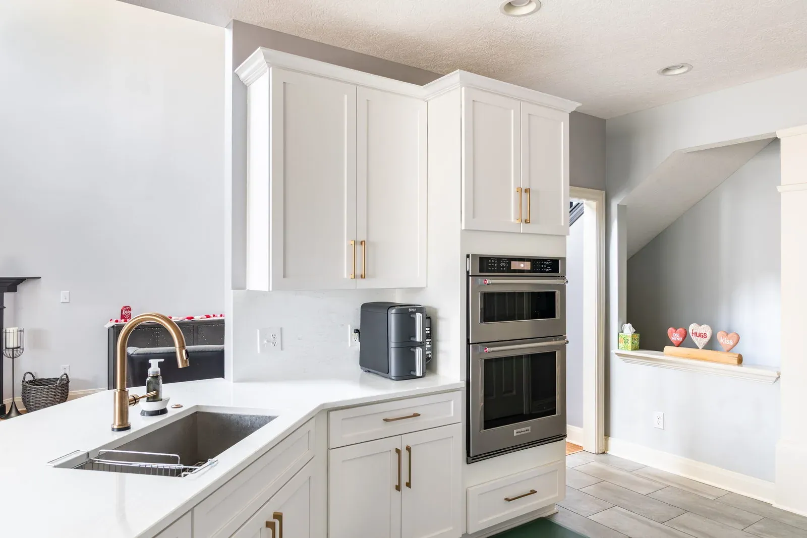 The same corner after remodeling, with a white shaker sink base, quartz countertops, a brass gooseneck faucet and stainless double wall ovens