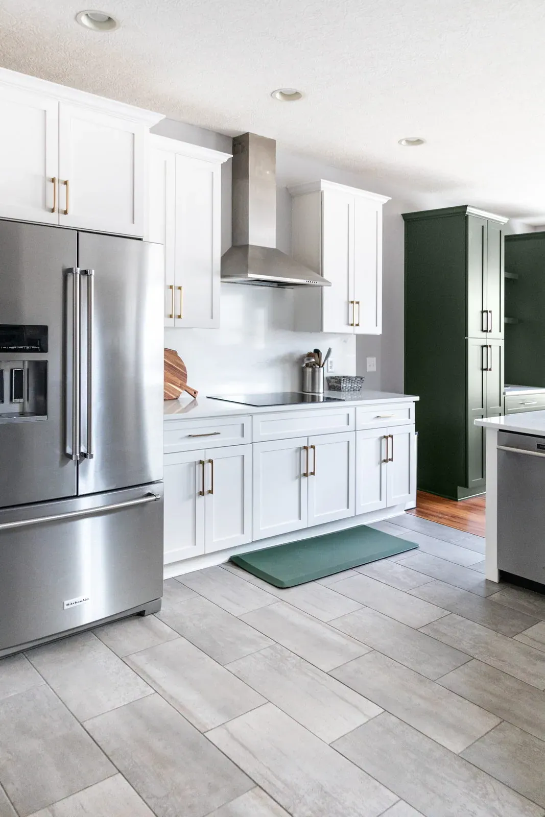 The same cooktop wall after remodeling, with white shaker cabinets, brass pulls, an induction cooktop under a stainless hood and grey plank tile floors