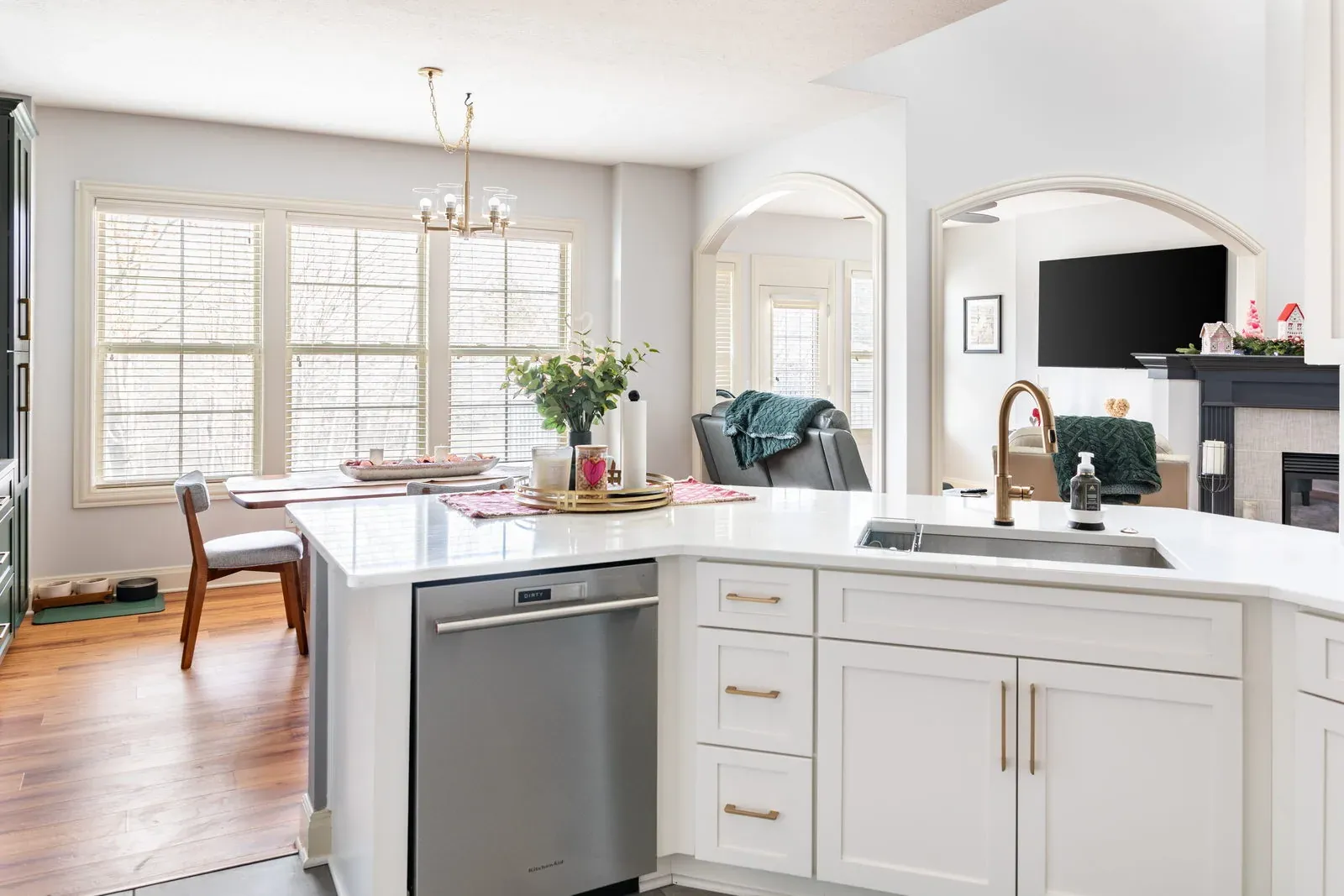 White shaker peninsula with a quartz top and brass faucet looking through an arched opening into a hearth room with a fireplace